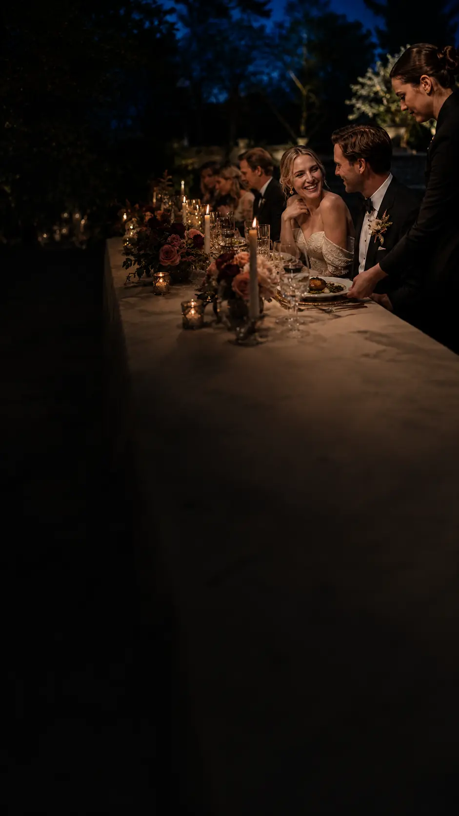 Newlyweds sharing a candlelit dinner as a catering professional serves their meal